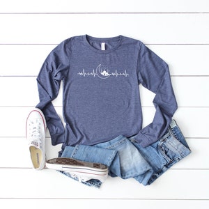 May include: A navy blue long-sleeved t-shirt featuring a white graphic of a crescent moon and a mosque with a heartbeat line. The shirt is laid flat on a white wooden surface with a pair of white trainers and blue jeans.