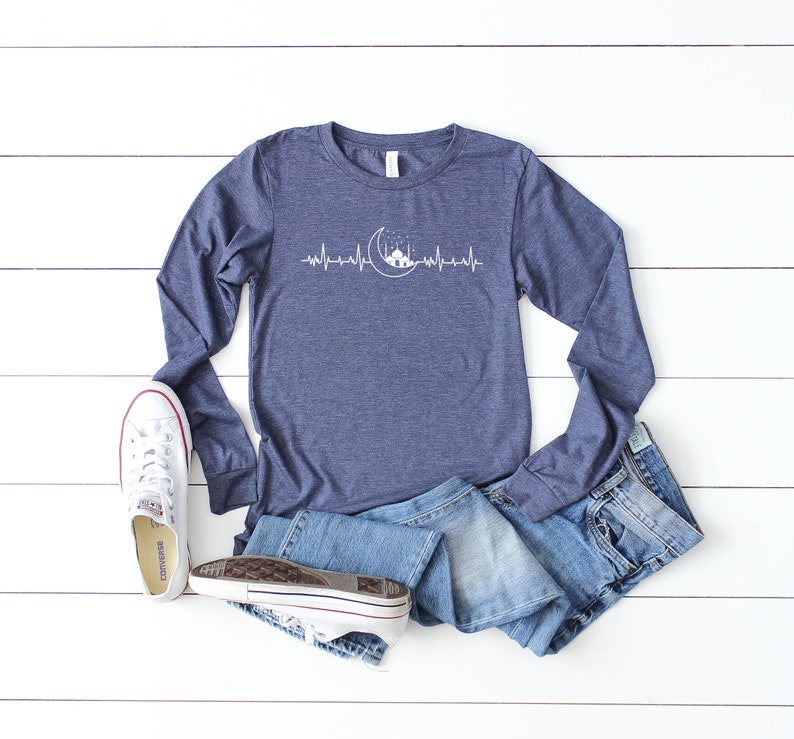 May include: A navy blue long-sleeved t-shirt featuring a white graphic of a crescent moon and a mosque with a heartbeat line. The shirt is laid flat on a white wooden surface with a pair of white trainers and blue jeans.