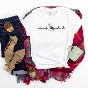 May include: White t-shirt featuring a black and white graphic of a crescent moon and mosque with a heartbeat line. The shirt is displayed on a red and black checked blanket with a pair of brown sandals, blue jeans, and a green mug.