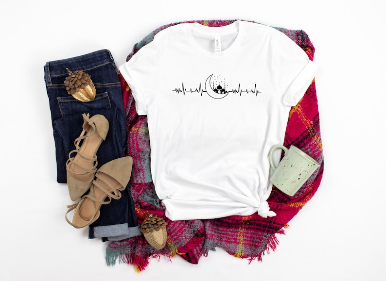 May include: White t-shirt featuring a black and white graphic of a crescent moon and mosque with a heartbeat line. The shirt is displayed on a red and black checked blanket with a pair of brown sandals, blue jeans, and a green mug.