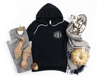 May include: Black hooded sweatshirt with a white monogram on the front pocket. The sweatshirt is laid out on a white surface with a pair of gray jeans, brown shoes, a white cotton branch, and a gold pumpkin.