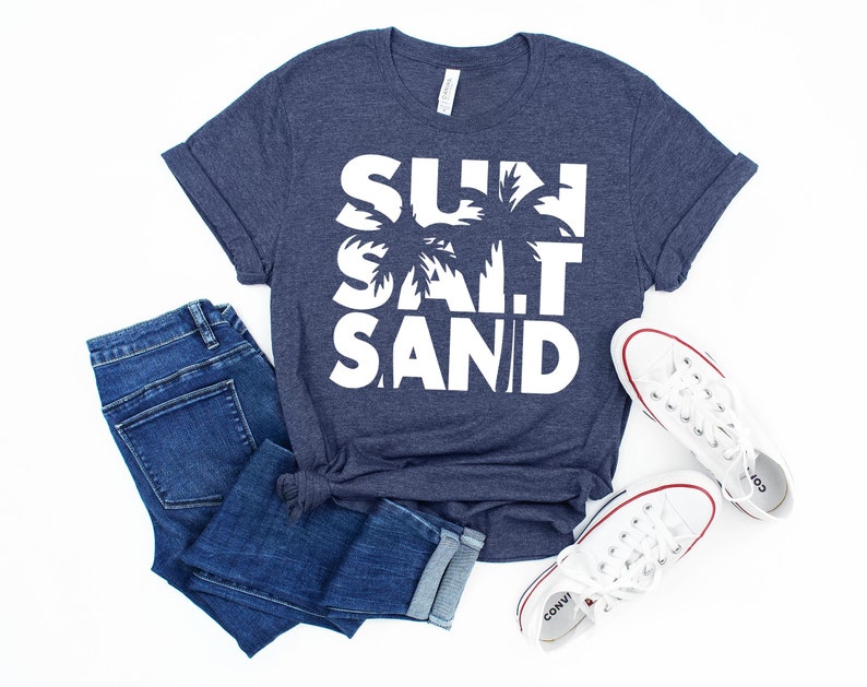 May include: A navy blue t-shirt with white text that reads "SUN SALT SAND" with a palm tree graphic. The shirt is paired with blue jeans and white sneakers.