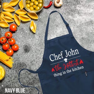 May include: Navy blue kitchen apron with a white pocket and the text "Chef John the Hottest thing in the kitchen" in white with red flames.