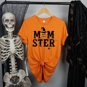May include: Orange t-shirt with a black graphic of a pumpkin wearing a witch hat and the text "MOMSTER" with a spider web and spider.
