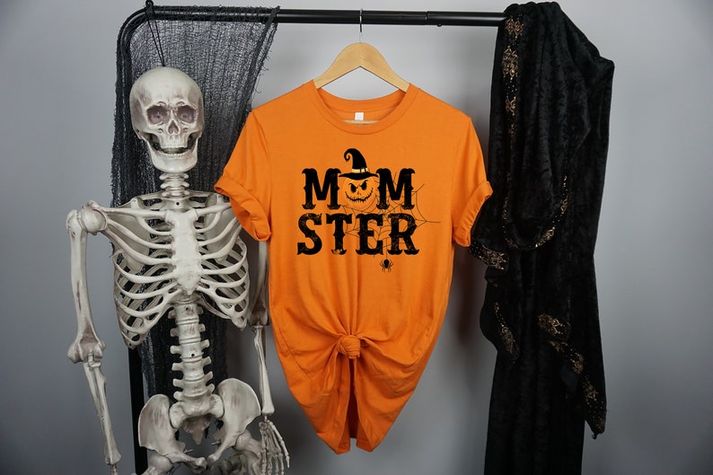 May include: Orange t-shirt with a black graphic of a pumpkin wearing a witch hat and the text "MOMSTER" with a spider web and spider.