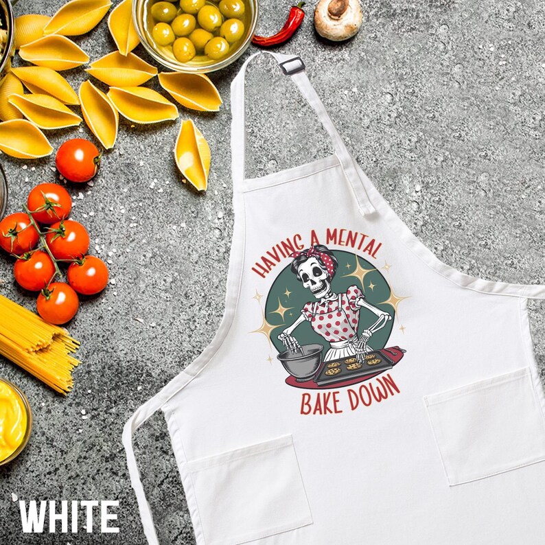 May include: White kitchen apron with a skeleton baking cookies. The apron has a pocket on each side and the text "Having a mental bake down".