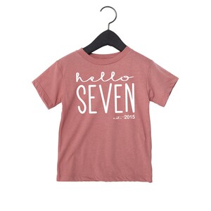 Peut inclure: Un t-shirt rose &agrave; manches courtes avec le texte "hello seven est. 2015" en blanc.