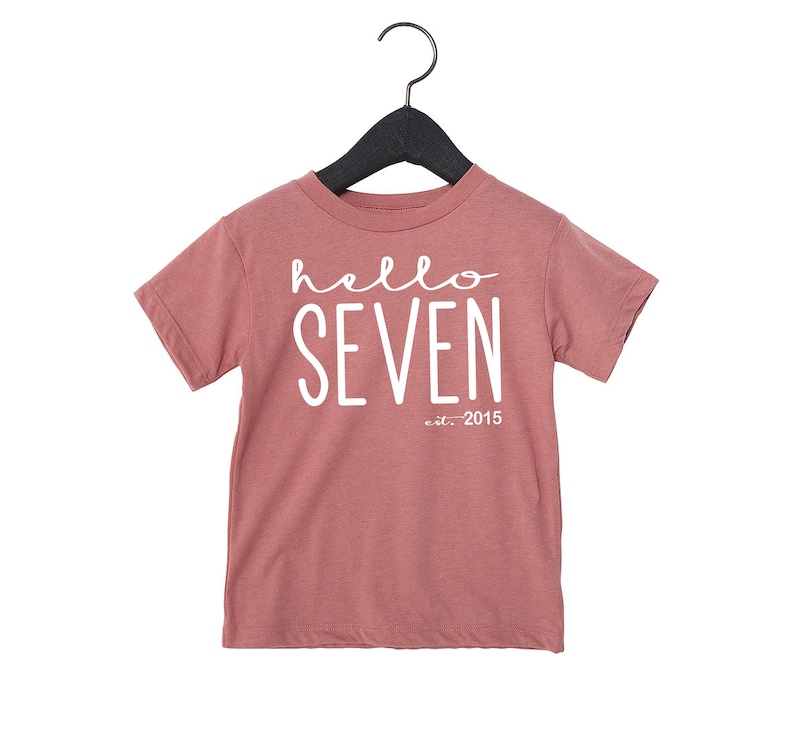 Peut inclure: Un t-shirt rose &agrave; manches courtes avec le texte "hello seven est. 2015" en blanc.