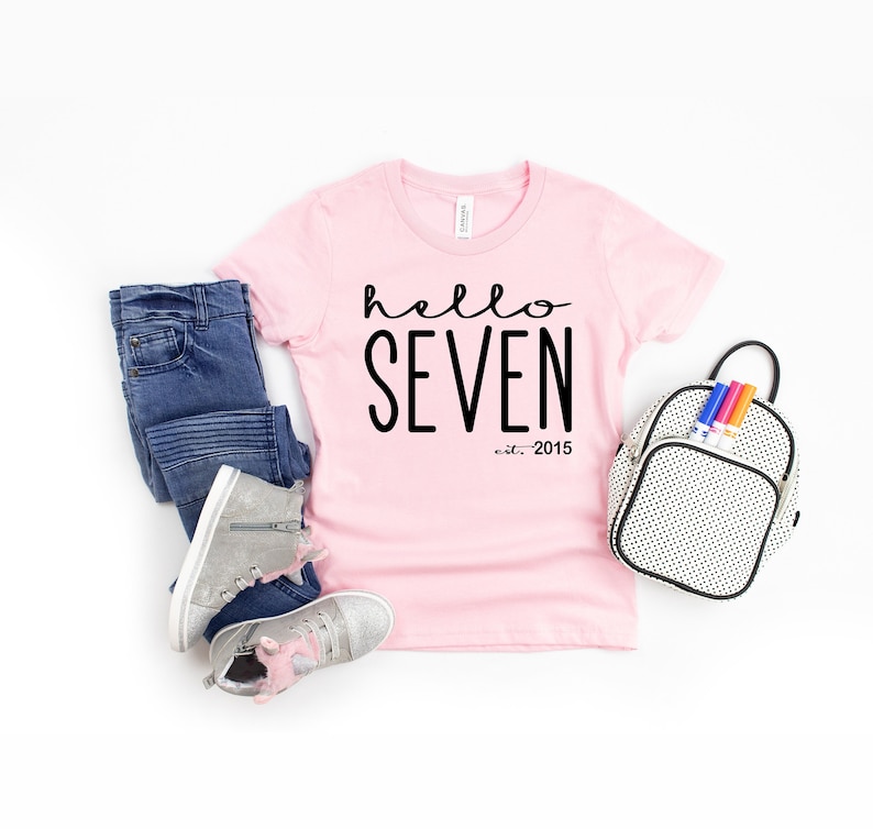 Peut inclure: T-shirt rose avec un texte noir qui dit "hello seven est. 2015".