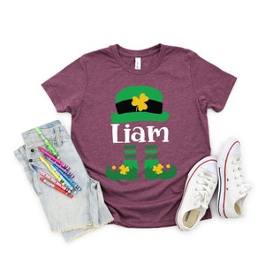 May include: A maroon t-shirt with a green leprechaun graphic and the name "Liam" printed on it. The leprechaun is wearing a green hat with a gold buckle and has green shoes with gold buckles.