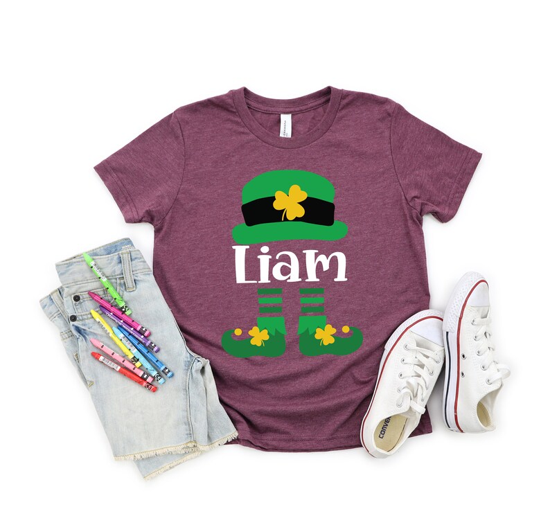 May include: A maroon t-shirt with a green leprechaun graphic and the name "Liam" printed on it. The leprechaun is wearing a green hat with a gold buckle and has green shoes with gold buckles.