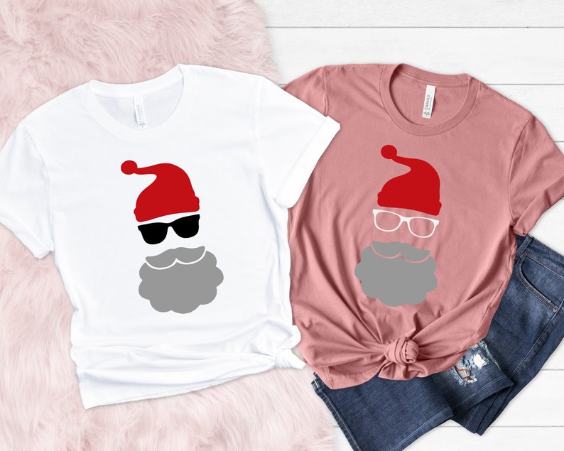 May include: Two t-shirts with a Santa Claus design. The white t-shirt has a black and gray Santa Claus face with sunglasses. The pink t-shirt has a white Santa Claus face with glasses.