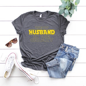 May include: Dark gray t-shirt with the text "BEST HUSBAND IN THE GALAXY" in yellow. The shirt is paired with blue jeans, white sneakers, and sunglasses. A sprig of greenery adds a decorative touch.