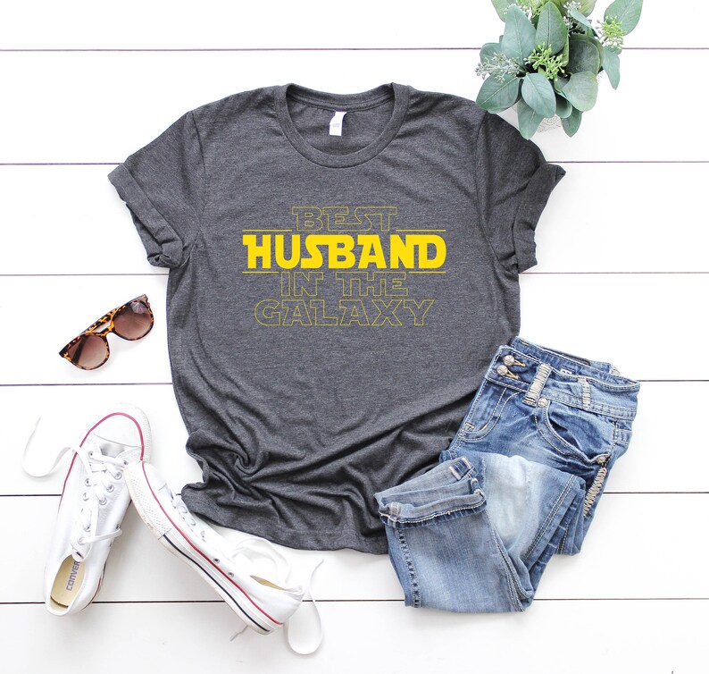 May include: Dark gray t-shirt with the text "BEST HUSBAND IN THE GALAXY" in yellow. The shirt is paired with blue jeans, white sneakers, and sunglasses. A sprig of greenery adds a decorative touch.