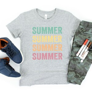 Puede incluir: Una camiseta gris con la palabra "SUMMER" repetida cuatro veces en diferentes colores. La camiseta se combina con pantalones de camuflaje, zapatillas azules de ca&ntilde;a alta y un juego de rotuladores de colores.