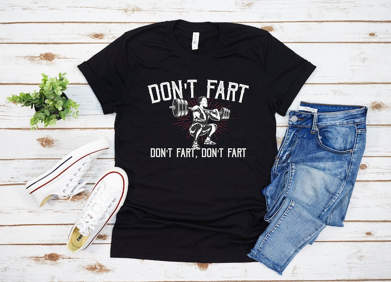 K&ouml;nnte beinhalten: Schwarzes T-Shirt mit einer wei&szlig;en Grafik einer Person, die Gewichte hebt. Der Text "Don't fart, don't fart" ist auf dem T-Shirt aufgedruckt.