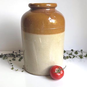 Może przedstawiać: Duży, dwutonowy ceramiczny słoik z zaokrągloną górą i cylindrycznym korpusem. Górna część jest w ciepłym brązie, a dolna w kremowej bieli. Czerwony pomidor spoczywa u podstawy. Słoik prawdopodobnie służy do przechowywania.