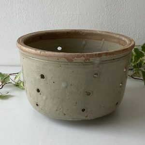 May include: A round, beige ceramic pot with numerous small holes and a reddish-brown rim. The pot has a rustic, aged appearance, suggesting it is vintage or antique. The design is simple and functional, suitable for various uses.