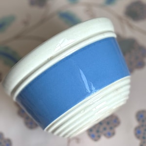 May include: A ceramic bowl featuring a smooth, glossy blue band around the center, contrasted by a cream-colored rim and a base with horizontal ribbed detailing. The bowl has a wide opening and a rounded shape, showcasing a two-tone color scheme.