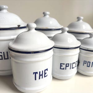 Puede incluir: Un conjunto de seis botes de cocina de esmalte blanco con tapas. Cada bote tiene un borde azul oscuro y una etiqueta azul oscuro con texto. Las etiquetas dicen "SU", "THE", "EPICE" y "POIVRE".