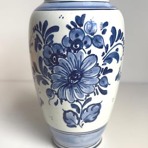 Può includere: Un vaso in ceramica bianca con motivi floreali dipinti a mano in blu. Il design presenta un grande fiore centrale circondato da fiori più piccoli, foglie e viticci. Il vaso ha un collo affusolato, un corpo arrotondato e bande blu.