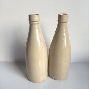 Puede incluir: Dos botellas cilíndricas altas hechas de cerámica esmaltada de color blanco roto. Cada botella presenta un cuello estrecho, un borde ligeramente ensanchado y un acabado liso y brillante. Las botellas están erguidas y tienen un color beige.
