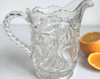 Puede incluir: Jarra de vidrio transparente con un patrón decorativo de flores y hojas. La jarra tiene un asa y un pico para verter. La jarra está al lado de una naranja en rodajas. La jarra probablemente se usa para servir bebidas.