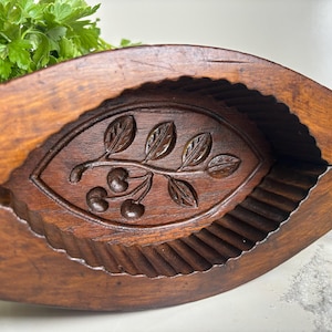 May include: A vintage wooden butter mould with a cherry and leaf design carved into the centre. The mould is a dark brown colour and has a scalloped edge. Fresh parsley is visible in the background.