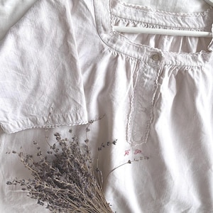 Puede incluir: Una blusa de lino en color beige claro de manga corta, con escote cuadrado adornado con encaje, una tapeta central con botones y pequeñas iniciales bordadas en rosa. Un manojo de tallos de lavanda seca se sitúa sobre la parte inferior.