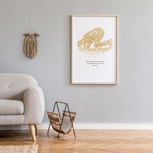 May include: A framed art print featuring gold calligraphy depicting a figure in prayer. The print is in a light wood frame and hangs on a gray wall. A beige sofa, a woven magazine rack, and a macrame wall hanging are also visible.