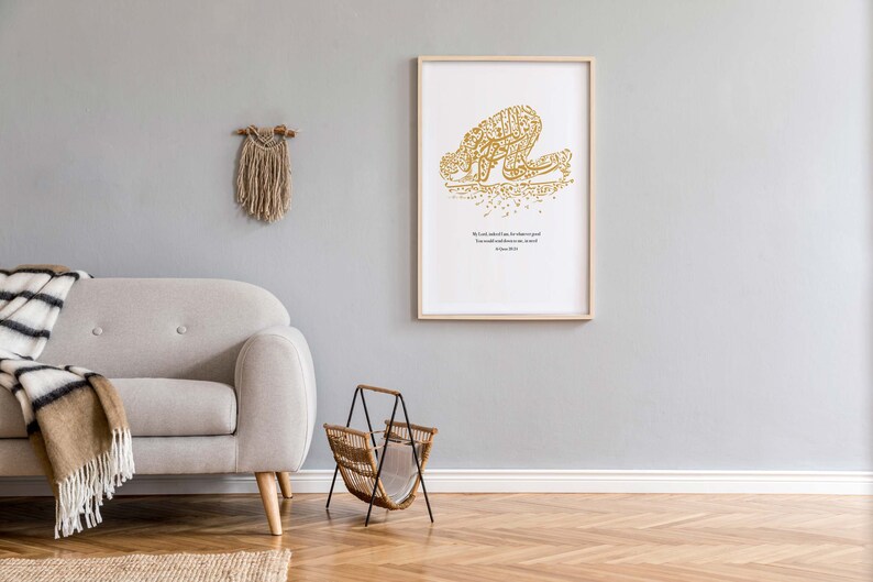 May include: A framed art print featuring gold calligraphy depicting a figure in prayer. The print is in a light wood frame and hangs on a gray wall. A beige sofa, a woven magazine rack, and a macrame wall hanging are also visible.