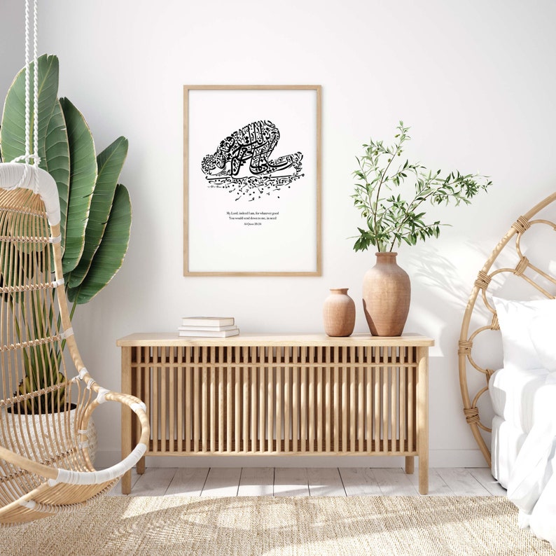 May include: A framed print featuring black Arabic calligraphy depicting a person in prayer. The print is in a light wood frame and includes English text. The print is displayed above a light wood cabinet with a natural woven chair and large green plant.