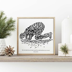 May include: A framed black and white calligraphy art print. The artwork features stylized Arabic script forming a kneeling figure. The text at the bottom reads "My Lord, indeed I am, for whatever good You would send down to me, in need".