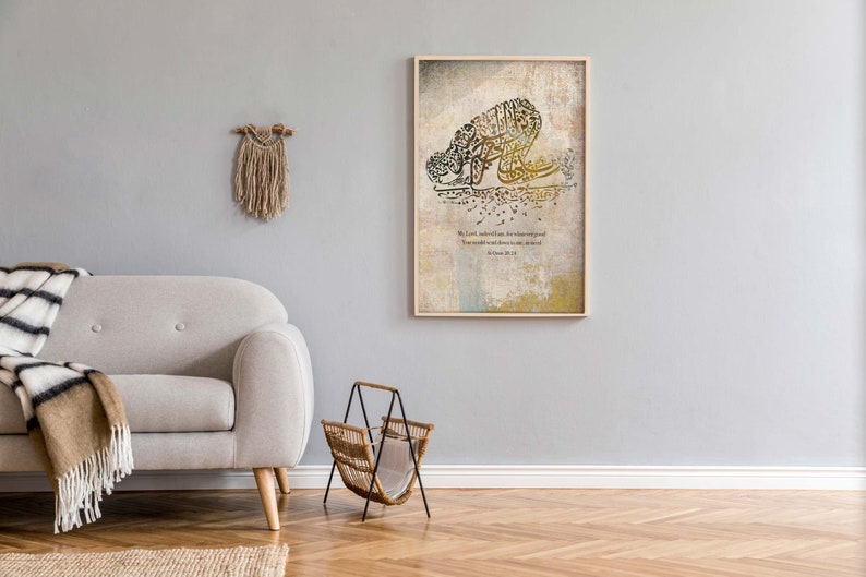 May include: A framed artwork featuring Arabic calligraphy in shades of gold and brown, depicting a figure in prayer. The artwork is displayed on a light gray wall, alongside a beige sofa, a woven magazine rack, and a decorative wall hanging.