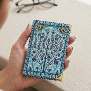 May include: A rectangular notebook with a tree design in blue, teal, and gold. The corners are reinforced with gold metal. The notebook is held in a hand.
