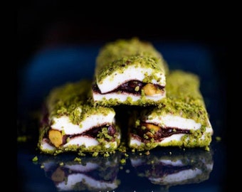 Puede incluir: Tres piezas de delicia turca cubierta de pistacho, cada una con una capa de chocolate y un relleno blanco. La delicia está apilada sobre una superficie azul oscuro.