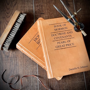 May include: Two leather-bound books, one titled "Book of Mormon" and the other titled "Doctrine and Covenants, Pearl of Great Price". The books are stacked on top of each other and have a brown leather cover. The text on the books is in gold lettering. The books are on a wooden surface.