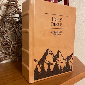以下が含まれることがあります： 表紙に「HOLY BIBLE KING JAMES VERSION」と刻印された茶色の革装丁の聖書。表紙には山と木のシルエットのデザインが施されています。