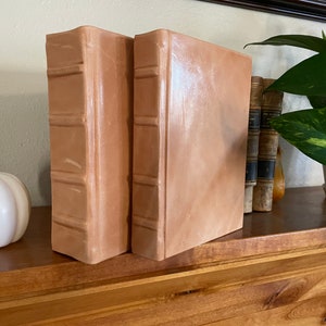 Op de afbeelding: Twee decoratieve boekenleggers gemaakt van bruin leer met een textuur. De boekenleggers staan op een houten plank met een kleine witte pompoen en een plant in een pot.