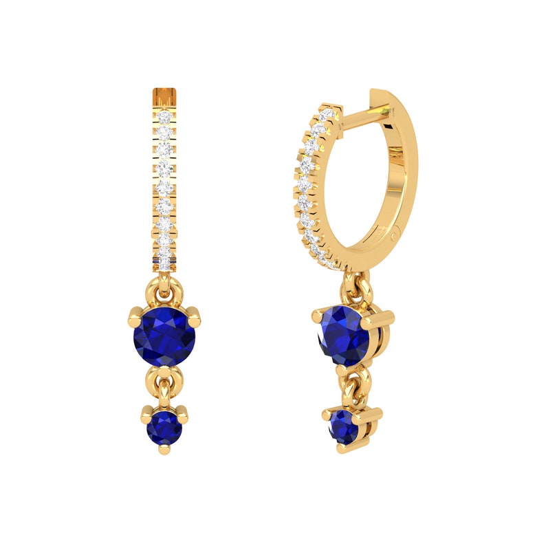 May include: Gold hoop earrings with a dangling charm featuring two blue sapphires. The hoop is accented with a row of small clear stones.