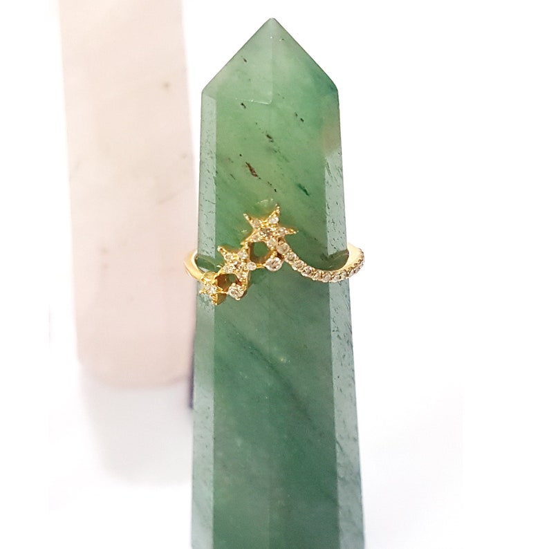 K&ouml;nnte beinhalten: Ein goldener Ring mit einem zarten Design aus drei Sternen, die mit kleinen, klaren Steinen besetzt sind. Der Ring wird auf einem gr&uuml;nen Stein pr&auml;sentiert.