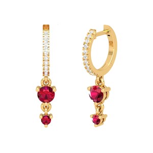 May include: Gold hoop earrings with a dangling charm featuring two red gemstones. The hoop is accented with a row of small clear gemstones.