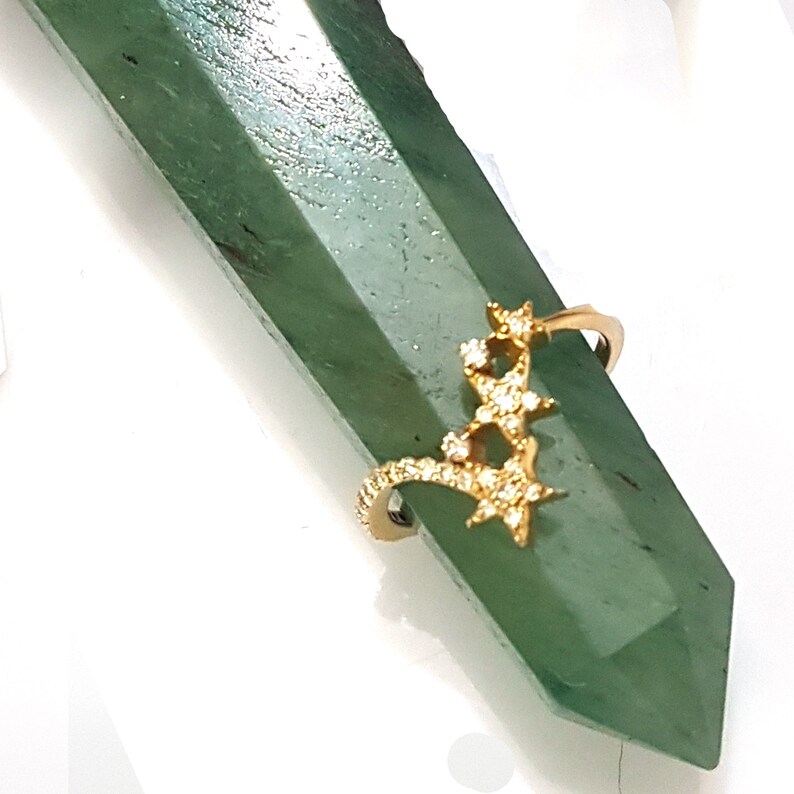 K&ouml;nnte beinhalten: Goldring mit einem Design aus f&uuml;nf Sternen, die mit kleinen, klaren Steinen besetzt sind. Der Ring liegt auf einem gr&uuml;nen Steinuntergrund.