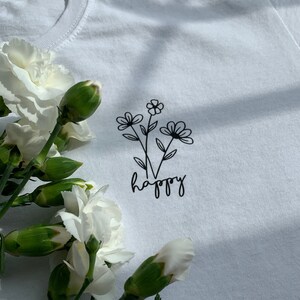 Könnte beinhalten: Weißes T-Shirt mit einer schwarzen Linienzeichnung von Blumen und dem Wort "happy" auf der Vorderseite.