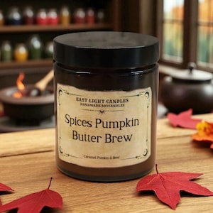 Könnte beinhalten: Eine Sojakerze in einem dunkelbraunen Glas mit schwarzem Deckel. Auf dem Etikett steht "Spices Pumpkin Butter Brew" von East Light Candles. Die Kerze steht auf einer Holzoberfläche mit roten und orangen Blättern.