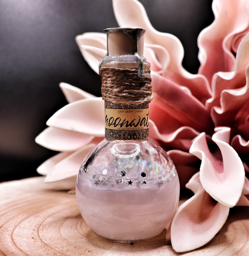 May include: A glass bottle with a cork stopper and a label that reads "Elixir Light Candy Moonwater". The bottle is filled with a clear liquid and has glitter and stars inside. The bottle is sitting on a wooden surface.