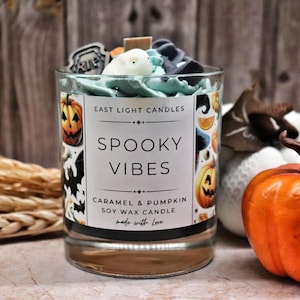 May include: A glass candle with a label reading "SPOOKY VIBES" and "CARAMEL & PUMPKIN", featuring pumpkin and bat illustrations. The candle has blue wax and decorative elements on top. An orange pumpkin is placed beside the candle.