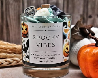 May include: A clear glass candle with a label that reads "SPOOKY VIBES" and "CARAMEL & PUMPKIN" with a pumpkin and bat design. The candle is topped with blue wax and decorative elements. A small orange pumpkin is next to the candle.