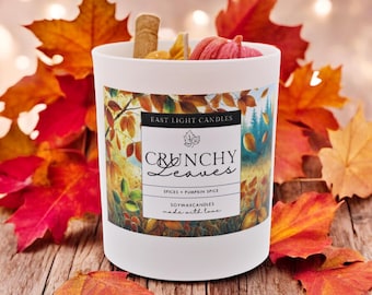 Könnte beinhalten: Eine weiße Kerze mit dem Text "Crunchy Leaves" und "Spices Pumpkin Spice" auf dem Etikett. Die Kerze ist mit einem Zimtstange und einem kleinen Kürbis verziert. Rote und orangefarbene Herbstblätter umgeben die Kerze.