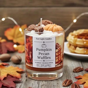 Könnte beinhalten: Ein Glas-Kerze mit der Aufschrift "Pumpkin Pecan Waffles". Die Kerze ist mit einem kleinen Kürbis und anderen Elementen verziert. Die Kerze ist von Herbstblättern und Pekannüssen umgeben.